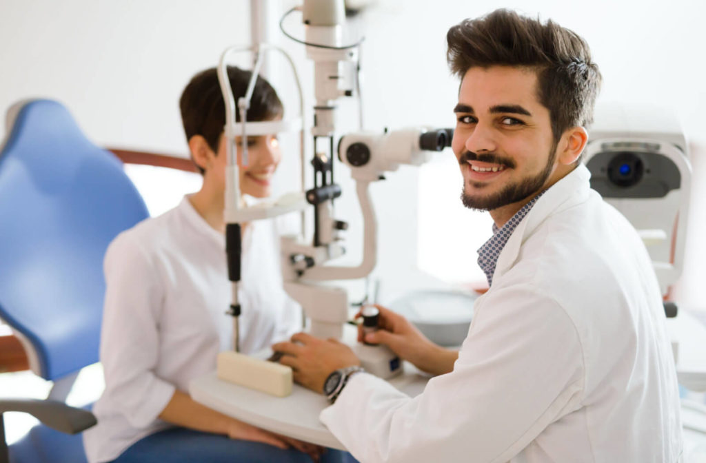 an optometrist looks behind him to smile at the camera, while a woman has an eye exam completed with a slit lamp