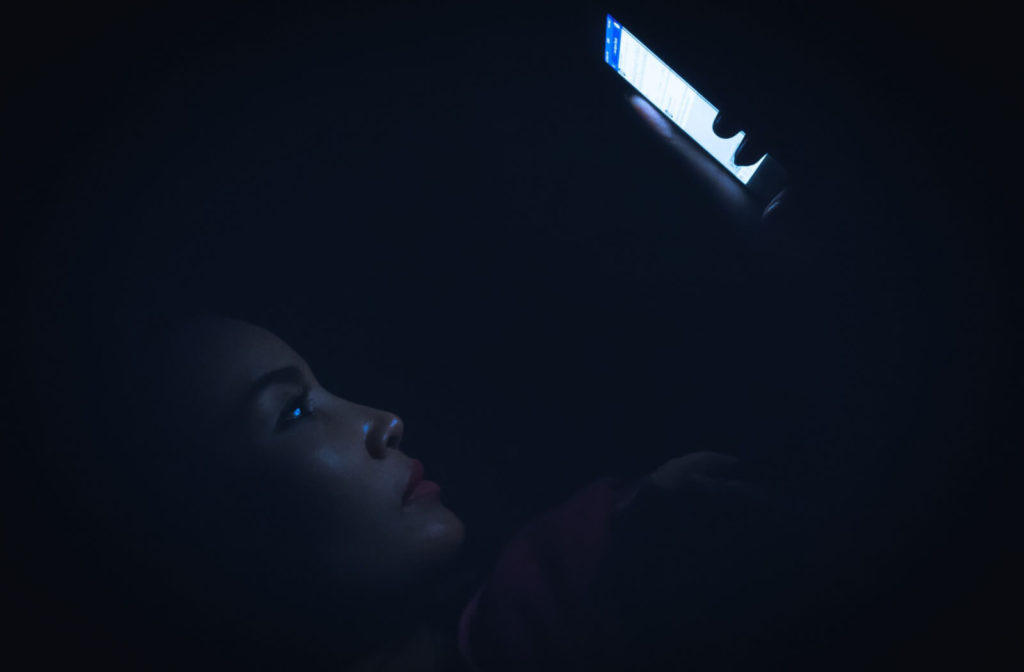 A young woman in bed browsing on her smartphone at late night.  blue light exposure in the evenings could be causing problems with a sleeping patterns which can contribute in dry eyes.