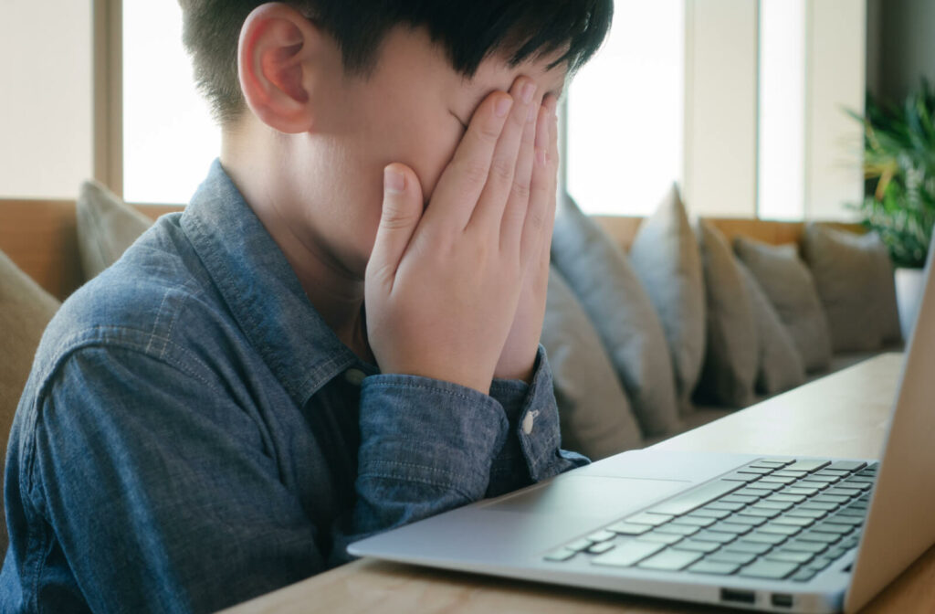 A young boy sitting in front of a laptop and covering his face with both hands