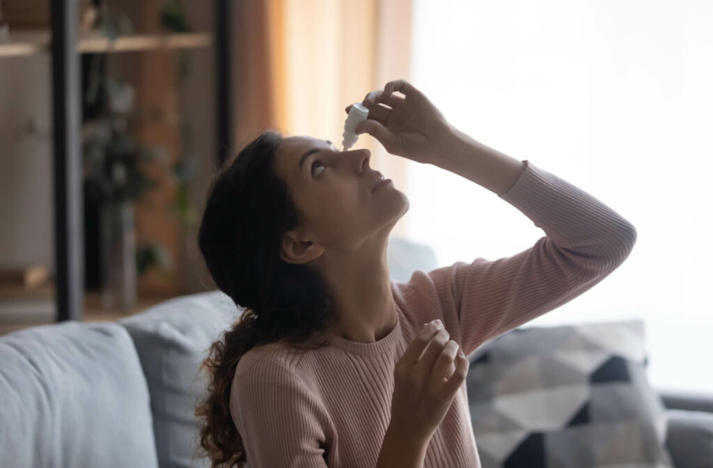 a  young woman sitting on a couch and tilting her head back as she applies eye drops to her left eye with her left hand