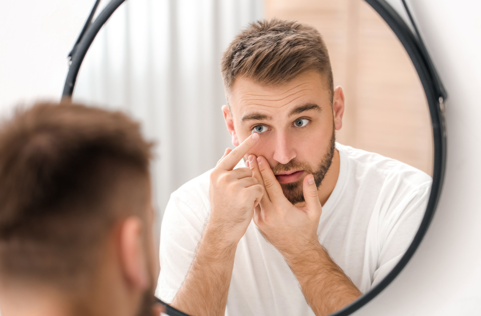 A young man looking into a mirror while he uses his left hand to put a contact lens in his left eye