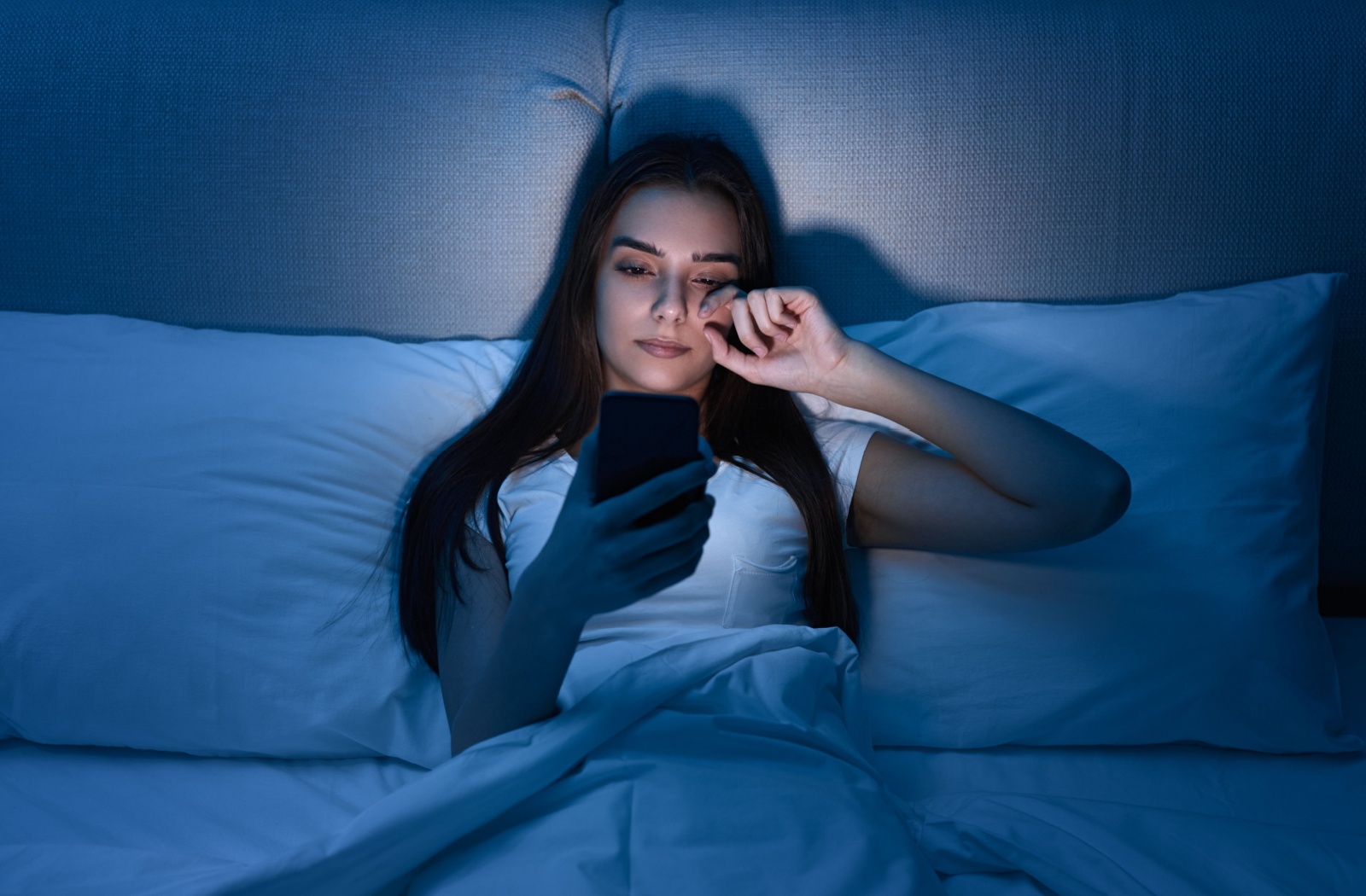 A young woman rubbing her eyes due to digital eye strain from using her cell phone in the dark.