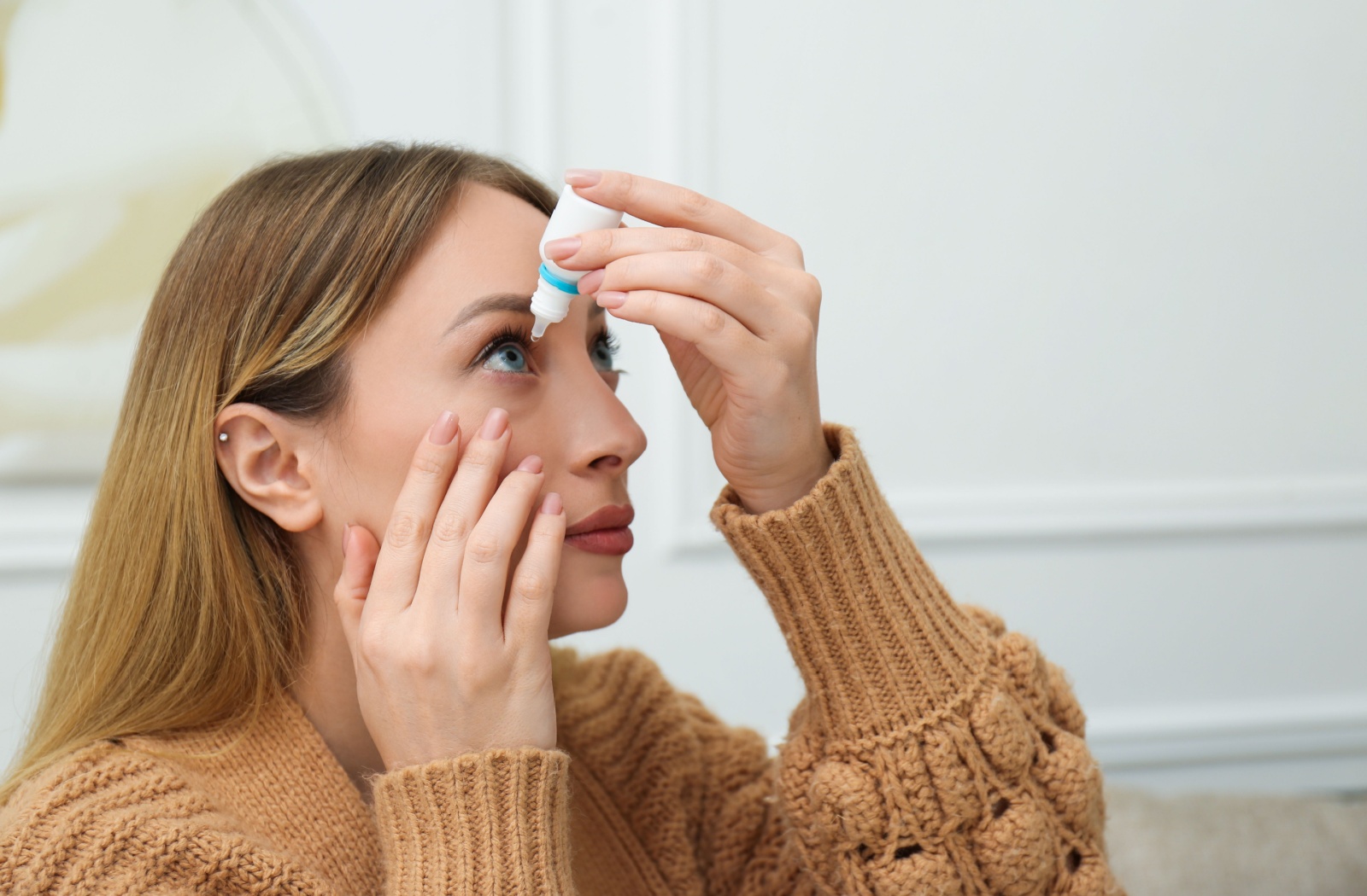 A person wearing a light brown chunky knit sweater applies eye drops in their right eye.