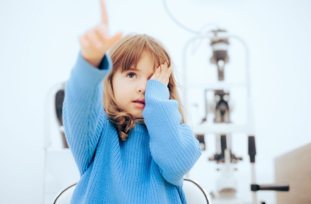 A young child in a blue sweater covering one eye and pointing out-of-frame during a children's eye exam.